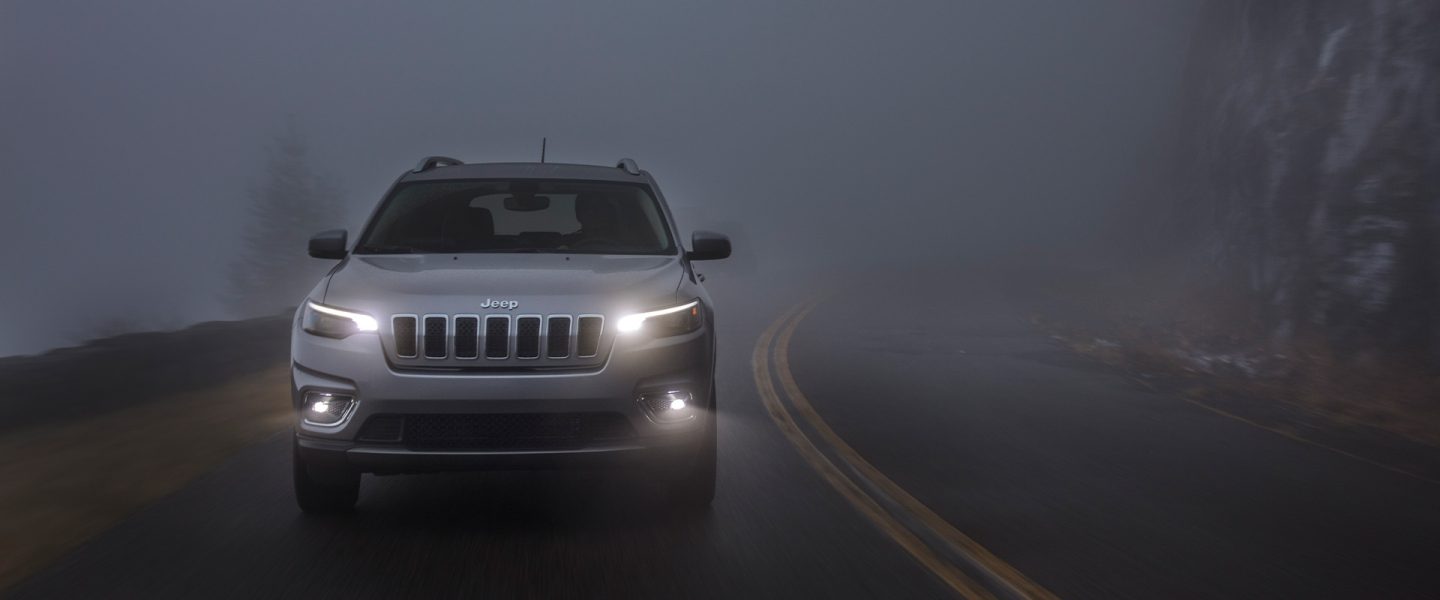 The 2020 Jeep Cherokee Limited on a foggy road at dusk with its headlamps and fog lamps lit.