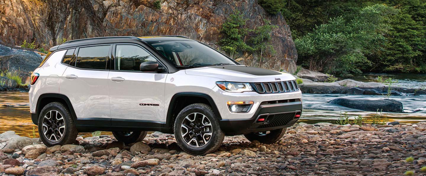 A white 2021 Jeep Compass Trailhawk parked on a rocky trail, next to a stream in the woods.